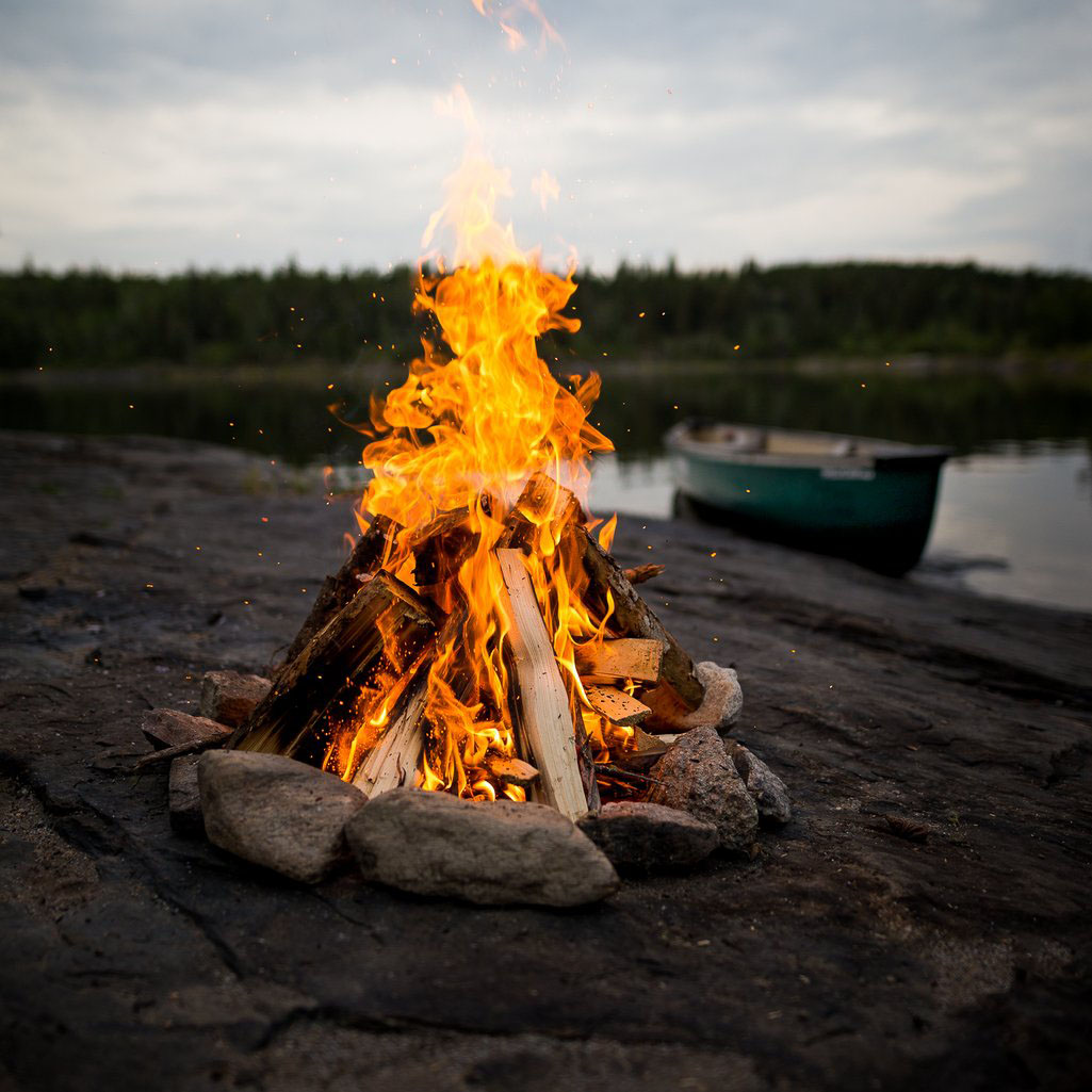 Campfire on the rocky shores of the Churchill River in Saskatchewan with a canoe in the background near the water.