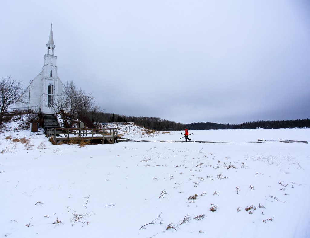 photo of person walking towards Holy Trinity Church near Stanley Mission holding cross-country skis on a gloomy winter day