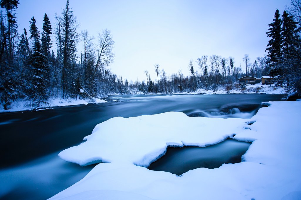 a long exposure photo of a river with snow covered shoreline and forest surrounding