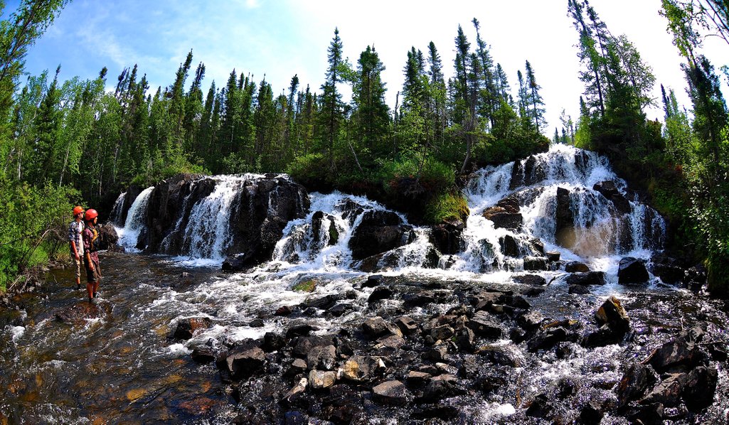 wide photo of two people standing at the base of a multi level waterfall in northern saskatchewan