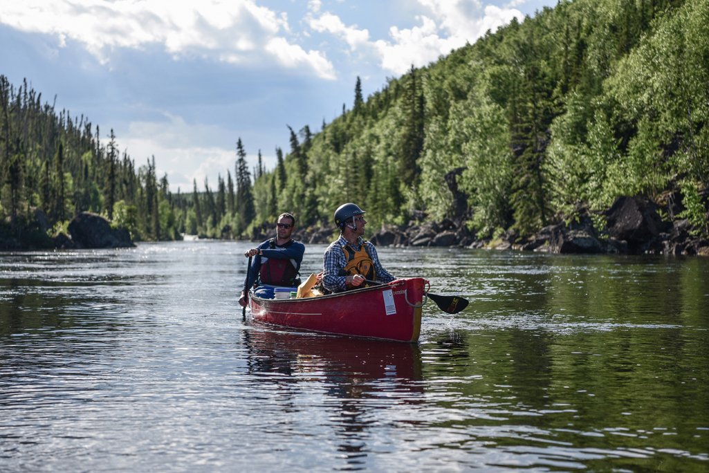 photo of a canoe with two paddlers in it going down a river with rocky shores and forest surrounding