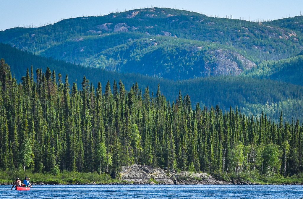 a small red canoe with two people in it paddling down a river with a large rocky hill or mountain in the distance