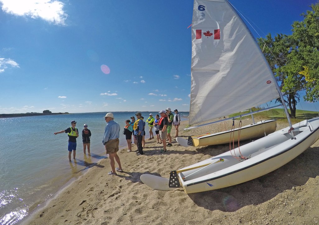 Sail boats on a beach beside a lake and people standing around listening to an instructor