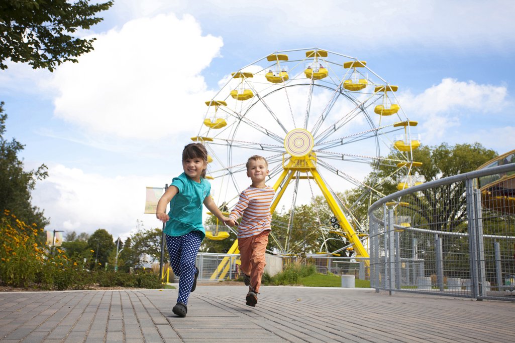 two young kids running and smiling with a ferris wheel in the background at Nutrien Playland in Saskatoon