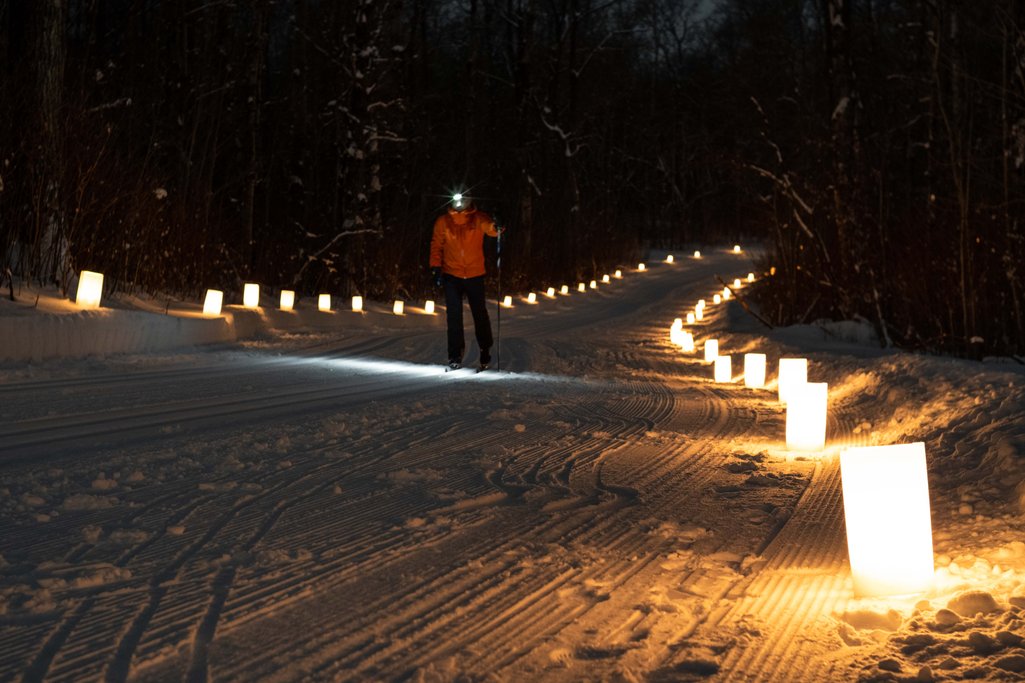 Person in red jacket cross-country skiing at night on a trail lined with candles on each side to illuminate the trail