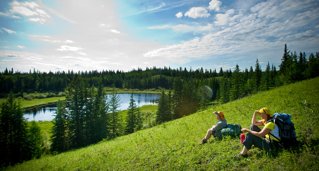 two hikers sitting on a hillside for a rest with backpacks on looking out to a lake and the boreal forest