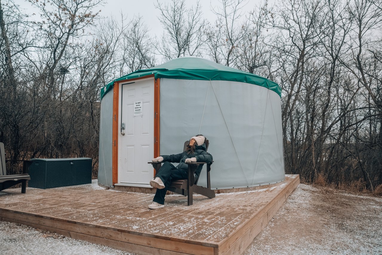 A woman sits in an Adirondak chair in front of a canvas yurt in winter with bear trees in the background.