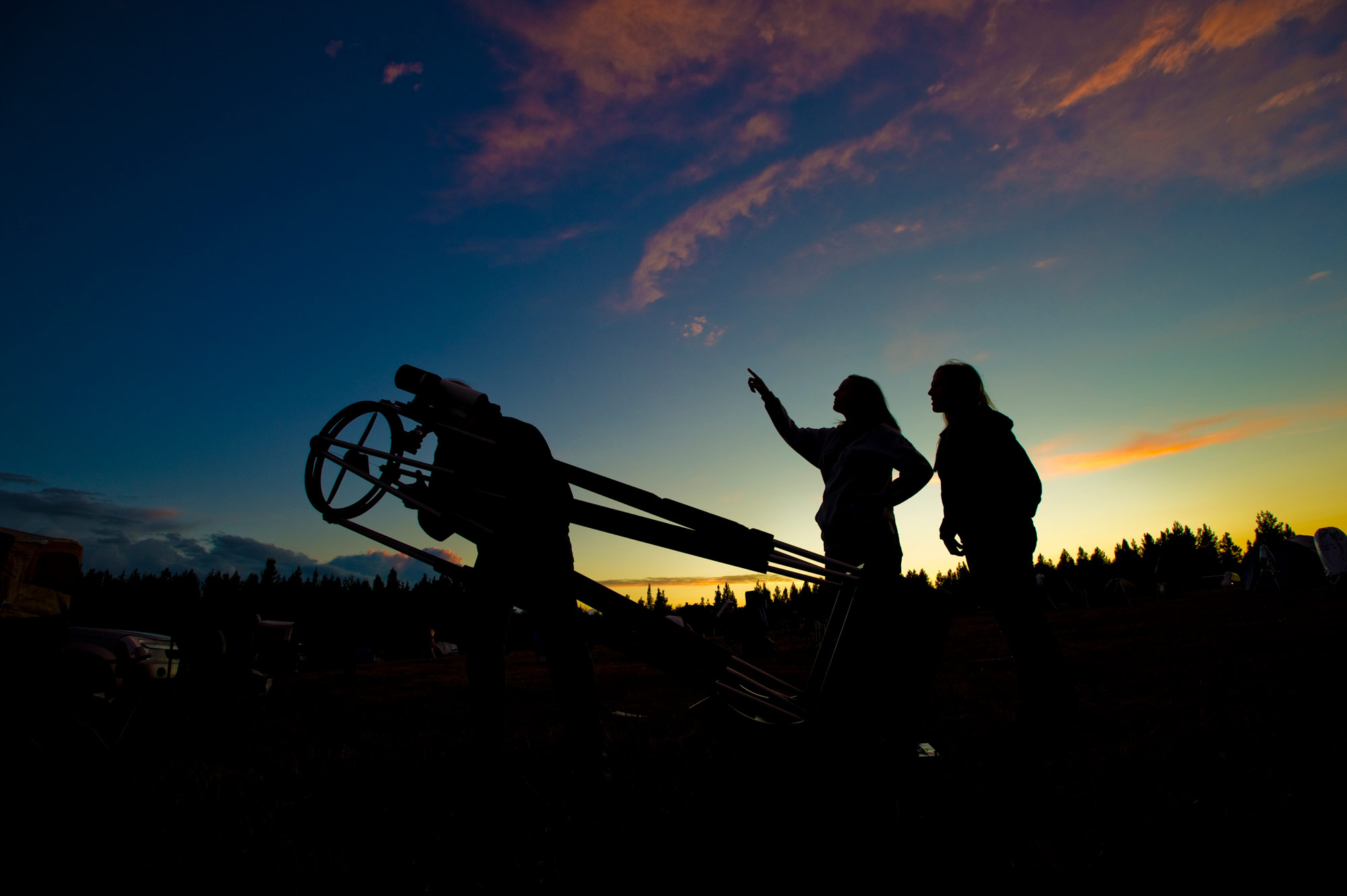 Three people looking through a telescope at the night sky with a sunset behind them