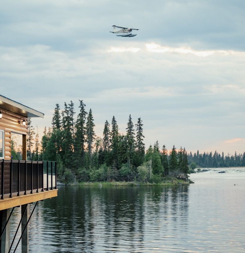 airplane flying over an island on water by wooden building, trees and mountains in distance